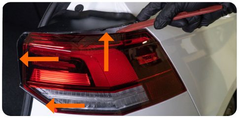 Pry tool at the outer tail light bezel of a VW Golf with three arrows marking the release points