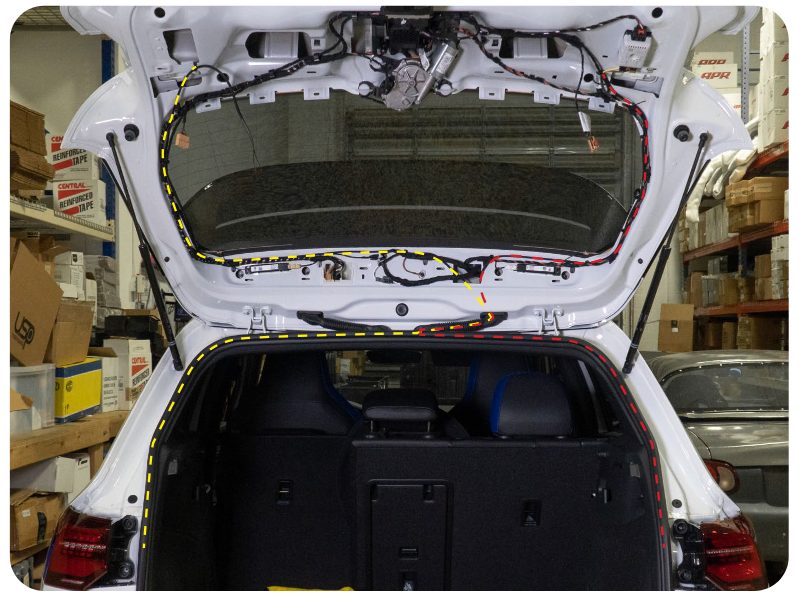 Rear of a VW Golf with dashed yellow and red lines tracing the harness route from the quarter panel tail lights up through the hatch to the inner tail lights